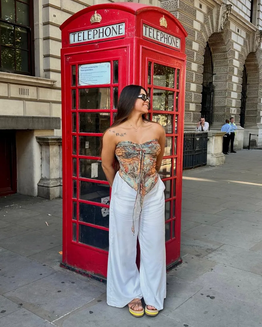 A Ester em pé na frente de uma cabine telefônica vermelha, numa rua de Londres.
